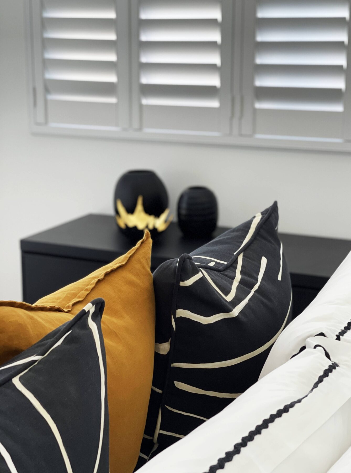 modern luxury black & white master bedroom with hint of mustard, gold decor on black console, white window shutters